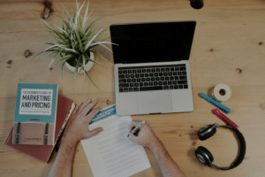 A workstation with a turned off laptop, notebooks, headphones. Someone's hands are in the photo looking like they are working.